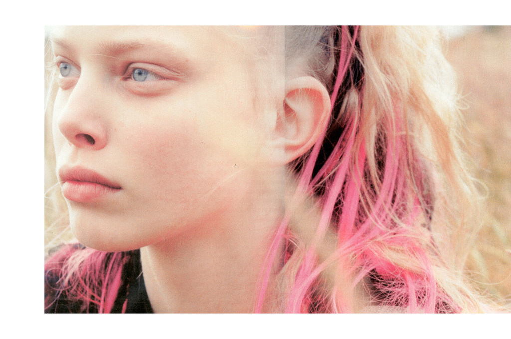 Close-up portrait of a young woman with fair skin, blonde hair with pink highlights, and blue eyes, set against a blurred background.