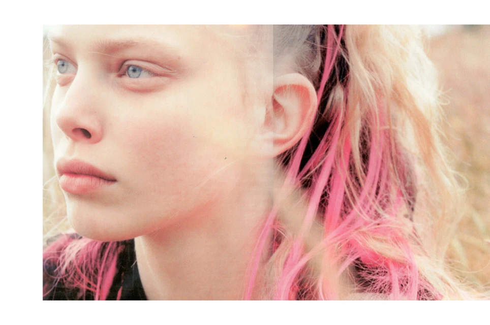 Close-up portrait of a young woman with fair skin, blonde hair with pink highlights, and blue eyes, set against a blurred background.