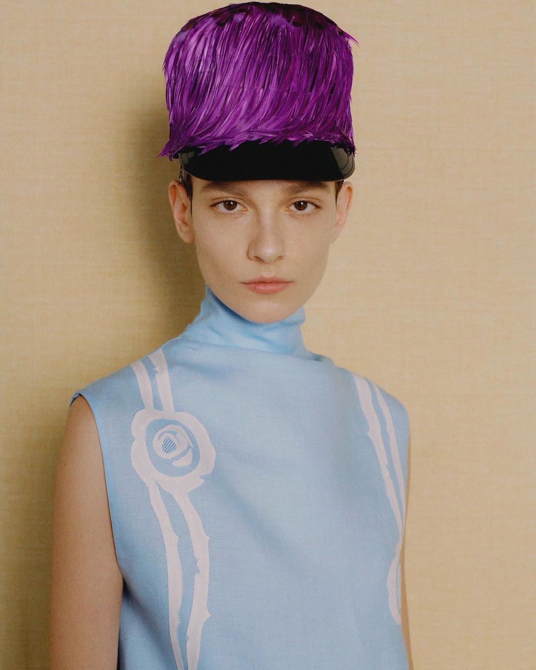 Portrait of a model wearing a light blue dress with white details and a purple feathered hat against a textured beige backdrop.