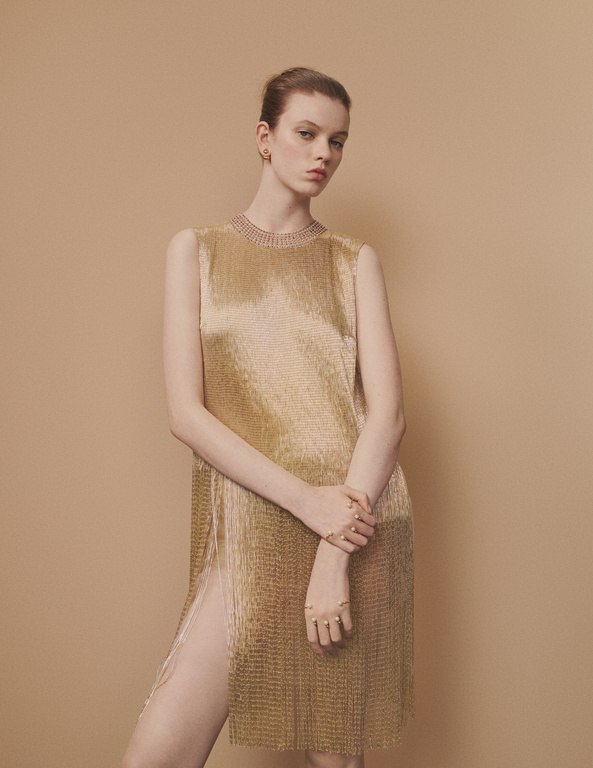 A slender woman with a serious expression poses in a golden, metallic, sleeveless, split dress against a neutral background in a studio shot.