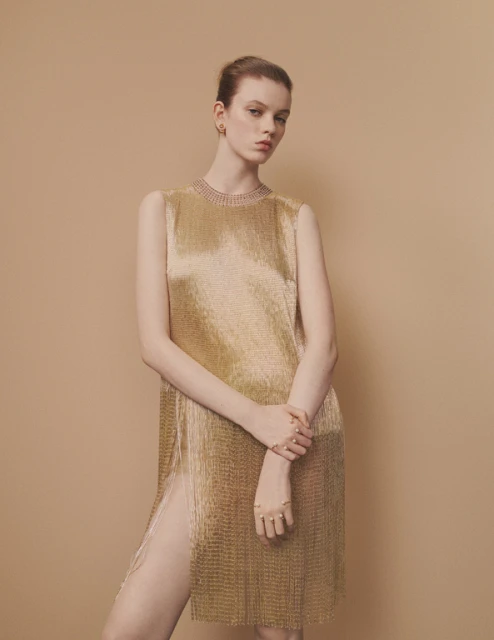 A slender woman with a serious expression poses in a golden, metallic, sleeveless, split dress against a neutral background in a studio shot.