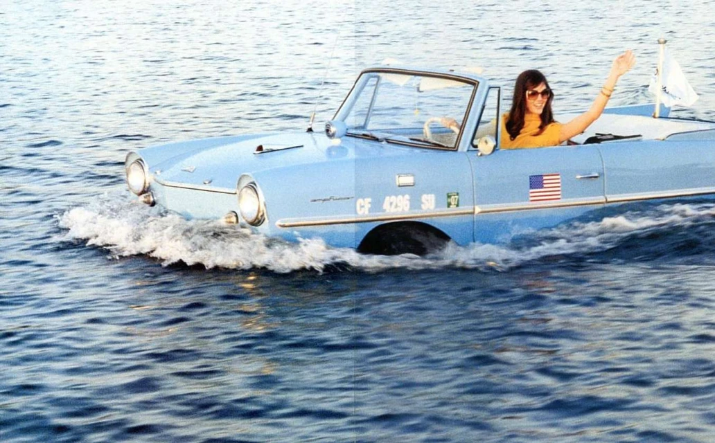 Light-blue Amphicar driving through blue water; woman in yellow shirt waves from the driver's seat of the vintage car, water splashing.