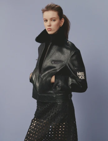 Young woman in a black leather jacket and skirt poses indoors with a medium shot on a blue background for a fashion editorial.