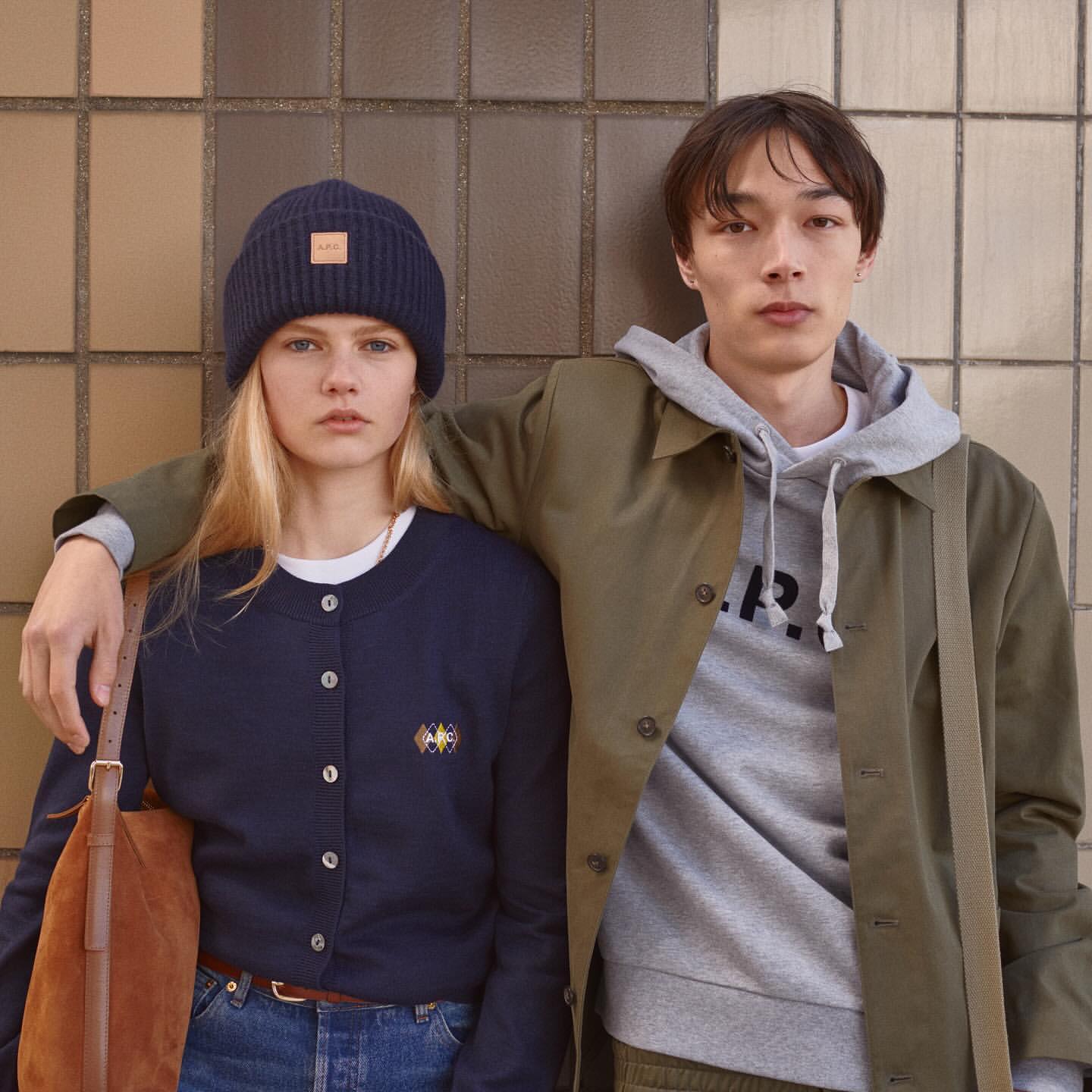 Two fashion models pose indoors against a tiled wall, wearing a navy beanie, cardigan, olive coat, and gray hoodie. Casual and cool style.