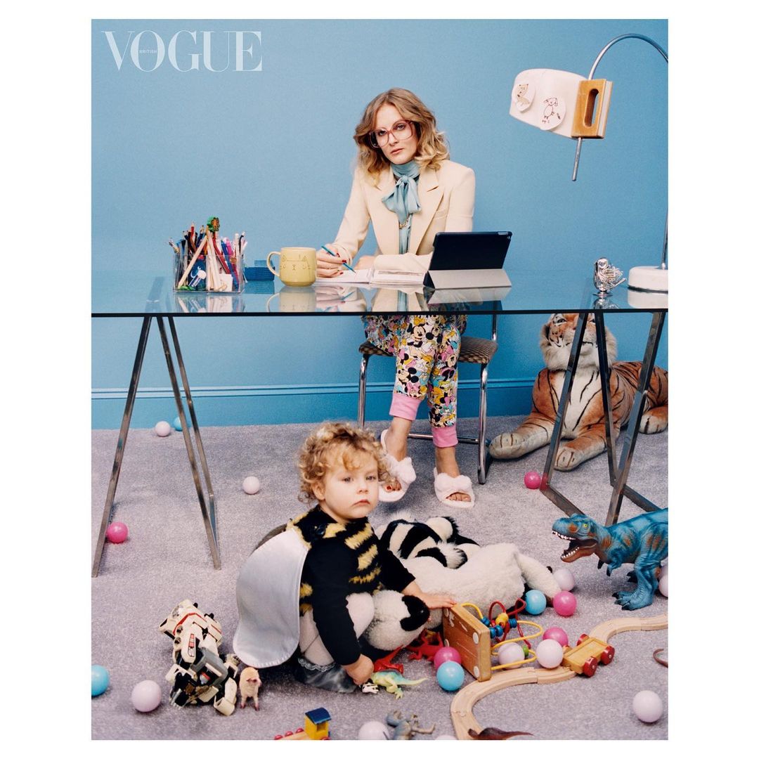 Woman works at a glass desk with a laptop and notebooks, while a child sits on the floor surrounded by toys in a blue room.