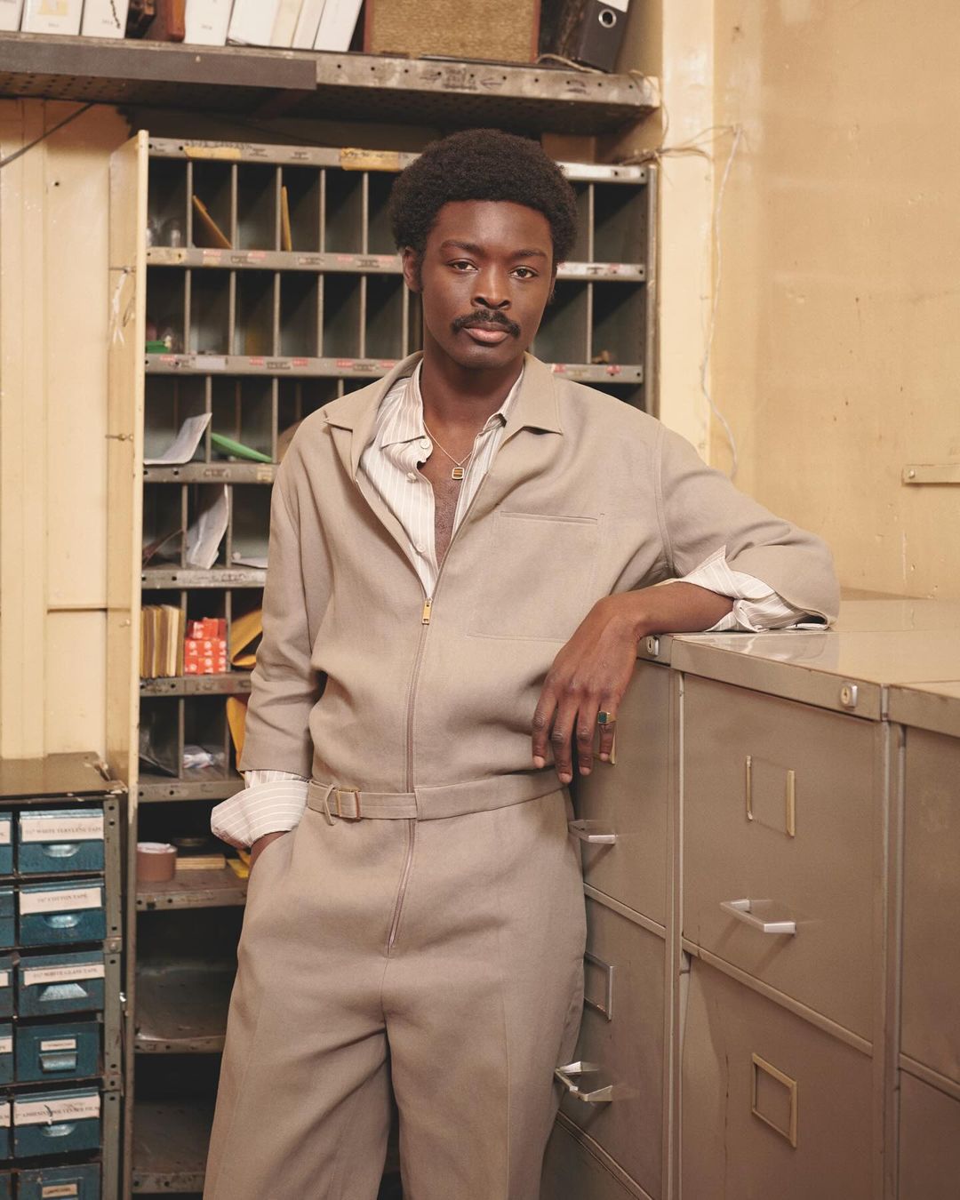 Stylish man in a beige jumpsuit poses in an indoor setting with filing cabinets and shelves, creating a retro, professional look.