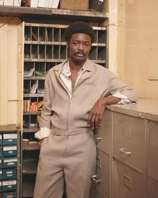 Stylish man in a beige jumpsuit poses in an indoor setting with filing cabinets and shelves, creating a retro, professional look.