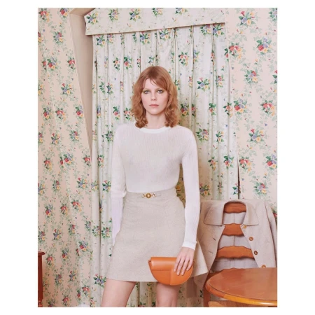 Indoor shot of a Caucasian woman in a white sweater and tan skirt, holding a purse, posing in front of floral wallpaper.