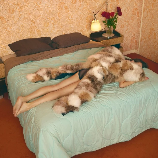 Woman lounging on a bed with a fur garment, complemented by a nightstand with a lamp and flowers against patterned wallpaper.