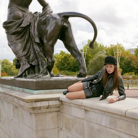 Adult female in a leather outfit poses lying on a stone ledge outdoors next to a large bronze statue and a background of trees and sky.
