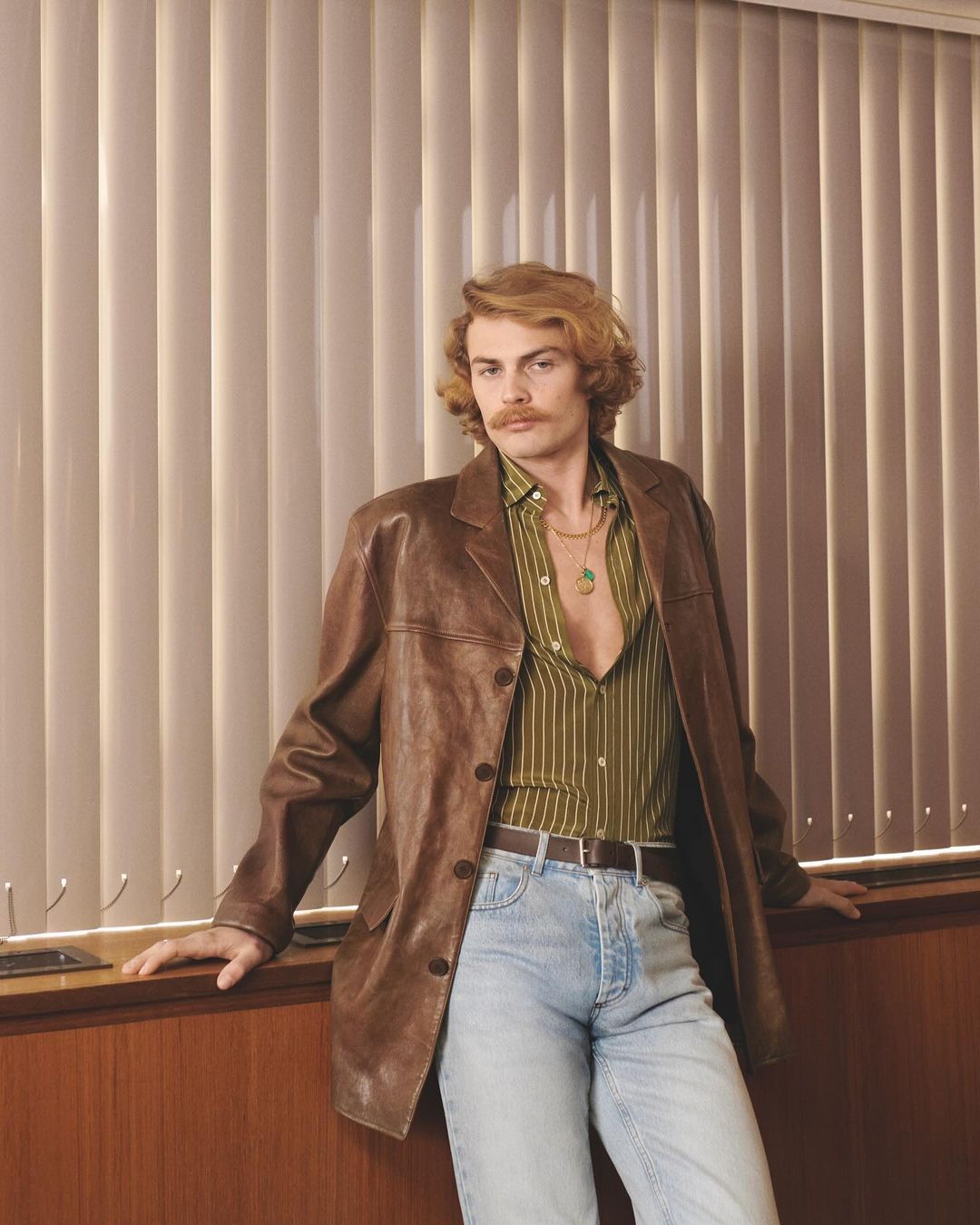 Stylish man in 70s-inspired fashion posing in front of window blinds, wearing a brown leather jacket, striped shirt, and light-wash jeans.