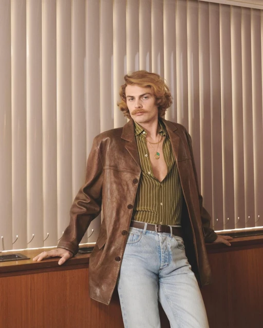 Stylish man in 70s-inspired fashion posing in front of window blinds, wearing a brown leather jacket, striped shirt, and light-wash jeans.