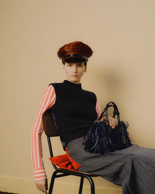 Fashion editorial shot of a woman in a black vest, striped top, and grey skirt, seated on a chair and holding a feathered handbag.