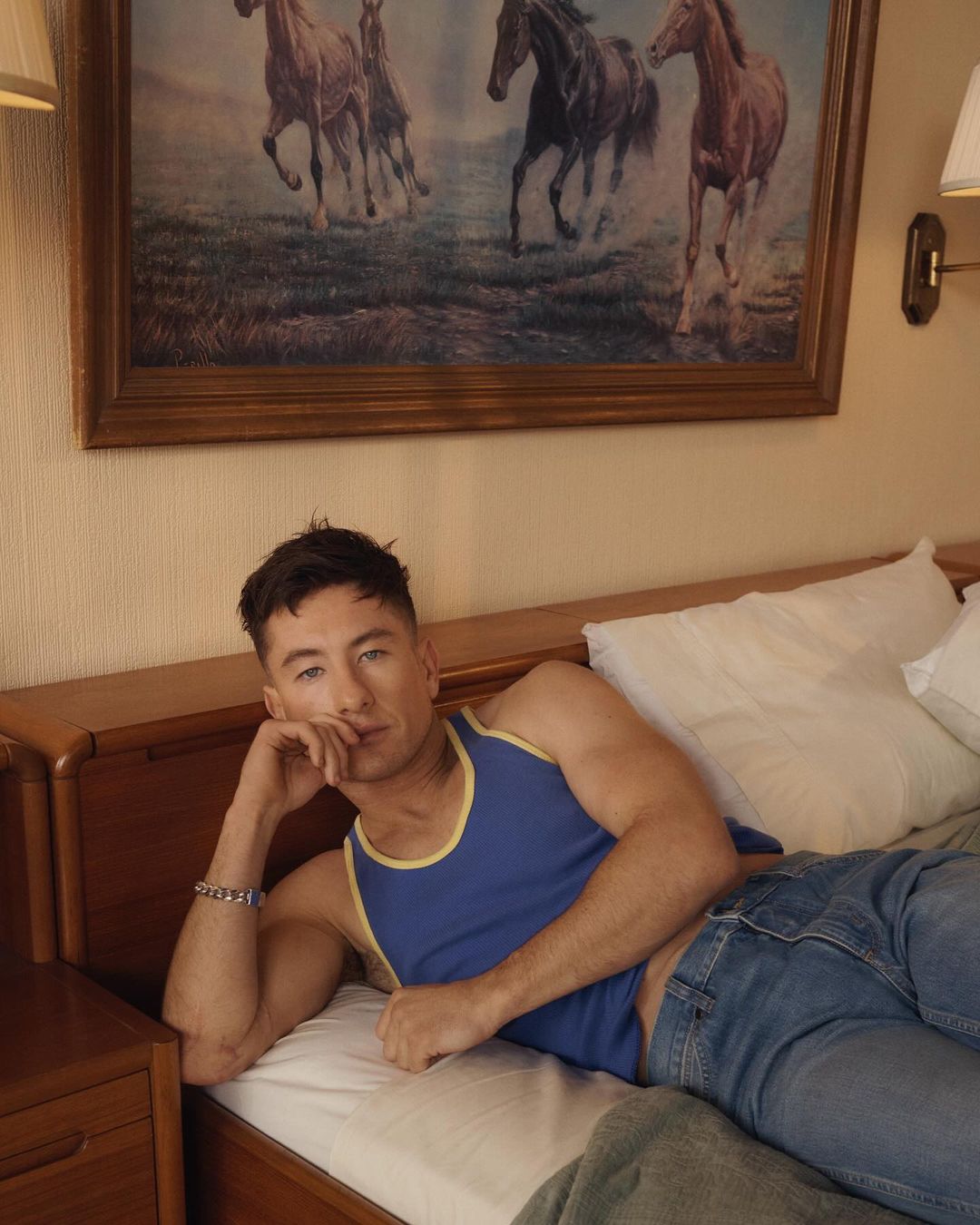 Actor Barry Keoghan poses while reclining on a bed in a tank top and jeans, with a painting of horses on the wall behind him.