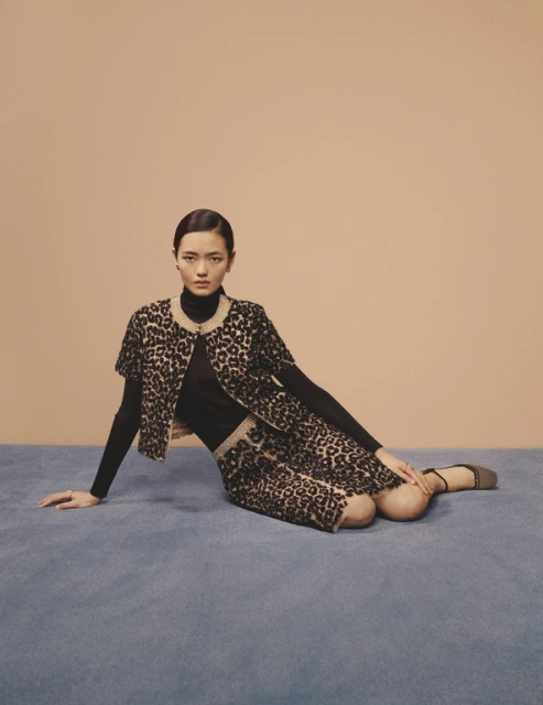 Fashion portrait of an Asian woman wearing a leopard print skirt and jacket with a black turtleneck on a blue surface.