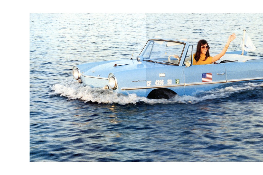 Light blue Amphicar convertible driving on water with a woman wearing sunglasses waving her hand and the American flag on the side.