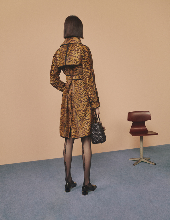 Woman in leopard-print coat stands indoors, facing away, holding handbag, wearing patterned tights, and shoes with small block heels.