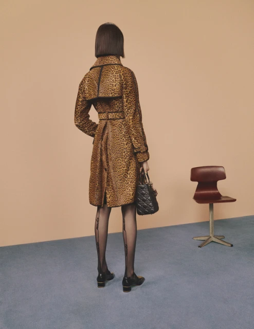 Woman in leopard-print coat stands indoors, facing away, holding handbag, wearing patterned tights, and shoes with small block heels.