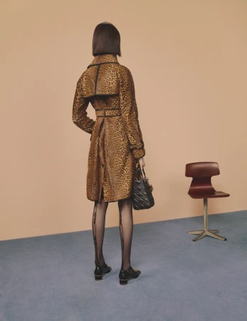 Woman in leopard-print coat stands indoors, facing away, holding handbag, wearing patterned tights, and shoes with small block heels.