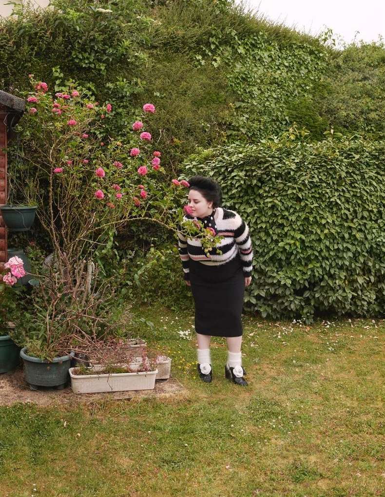 An adult female in a striped sweater stands in a green backyard, smelling pink roses growing on a bush. Potted plants sit in front.