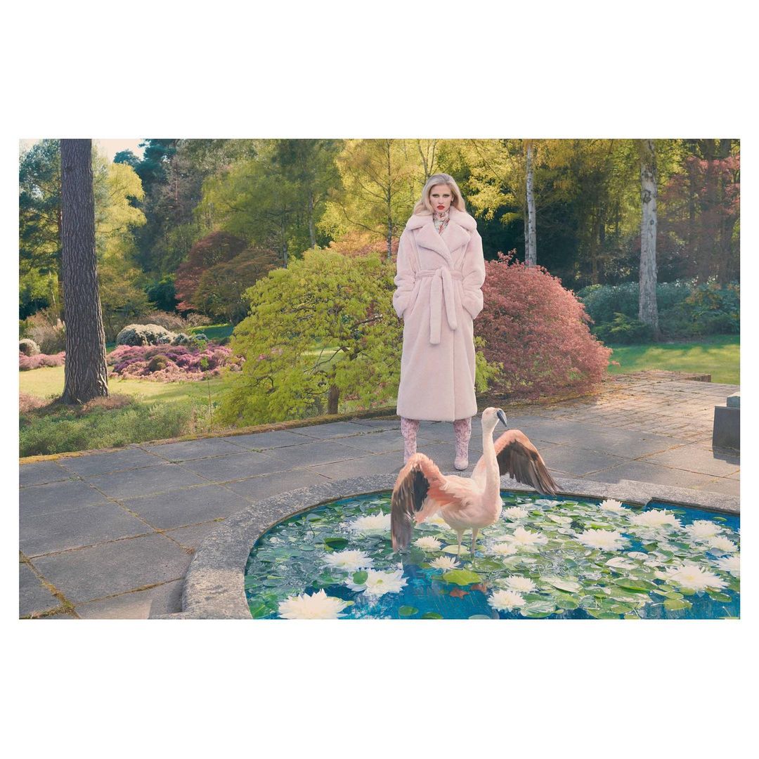 A woman in a pink coat stands near a pond with a flamingo, surrounded by a lush green landscape with trees and plants in soft light.