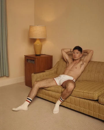 Man in white shorts and socks lounges shirtless on retro sofa, illuminated by a table lamp, posing casually in a neutral-toned interior.