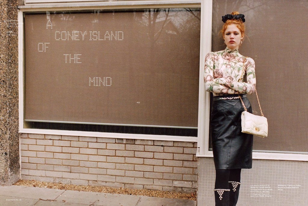 Fashion shot of an adult woman with red hair in a floral top and black skirt standing in front of a brick and concrete building.