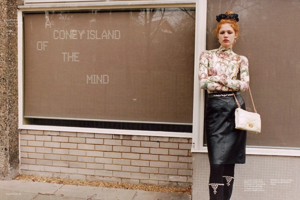 Fashion shot of an adult woman with red hair in a floral top and black skirt standing in front of a brick and concrete building.