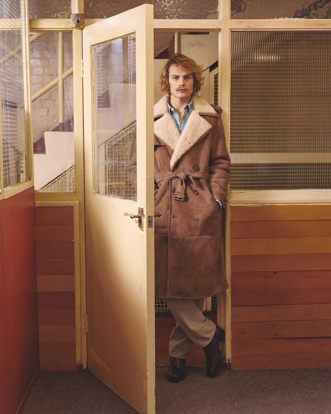 An adult male stands inside a doorway wearing a long brown coat, light pants, and black shoes in an indoor environment with light. 