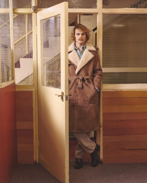 An adult male stands inside a doorway wearing a long brown coat, light pants, and black shoes in an indoor environment with light. 