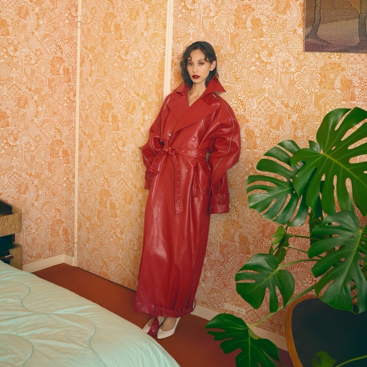 Elegant woman in a long red coat poses indoors against patterned wallpaper near a leafy green Monstera plant in an eye-level shot.
