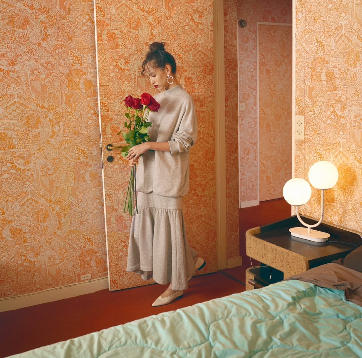 Indoor studio shot of a woman in casual clothing holding red roses, standing in front of wallpaper, with a bed and lamp visible.