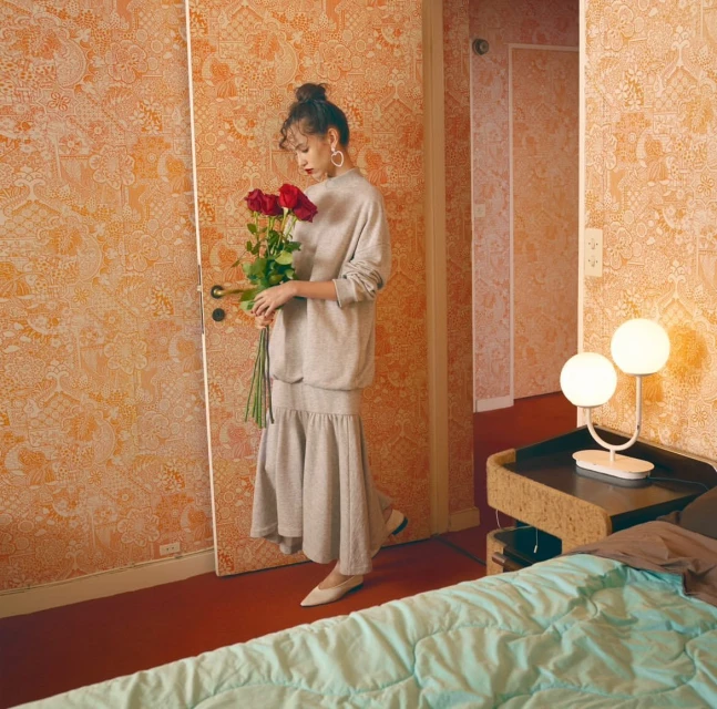 Indoor studio shot of a woman in casual clothing holding red roses, standing in front of wallpaper, with a bed and lamp visible.