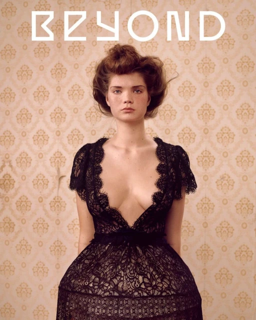 Portrait of a woman with auburn hair wearing a low-cut, black lace dress, standing in front of a vintage-patterned wall with the word 'BEYOND' above.