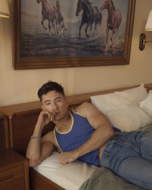 Adult male fashion model poses on a bed in jeans and blue tank top in an indoor setting. Horse painting on wall behind him.