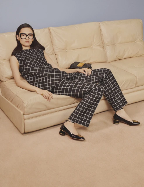 Adult woman in black-and-white checkered sleeveless top and pants lying on a tan couch in a minimalist studio.