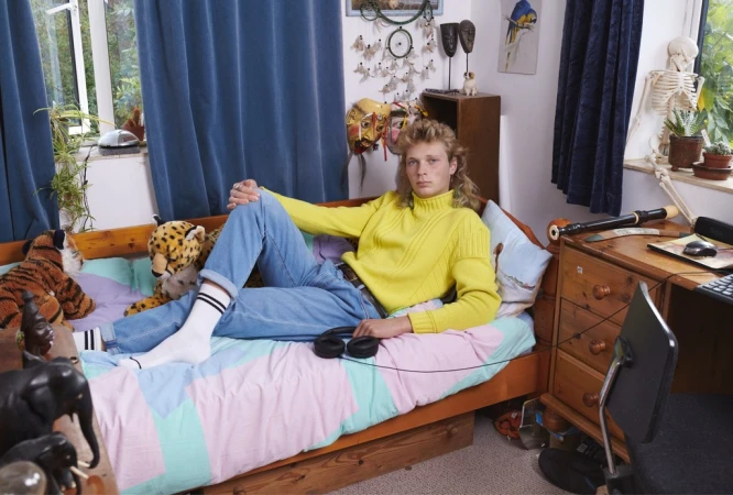Young adult relaxes on a bed with a yellow sweater, jeans, and white socks, surrounded by plush toys in a home interior with natural light.