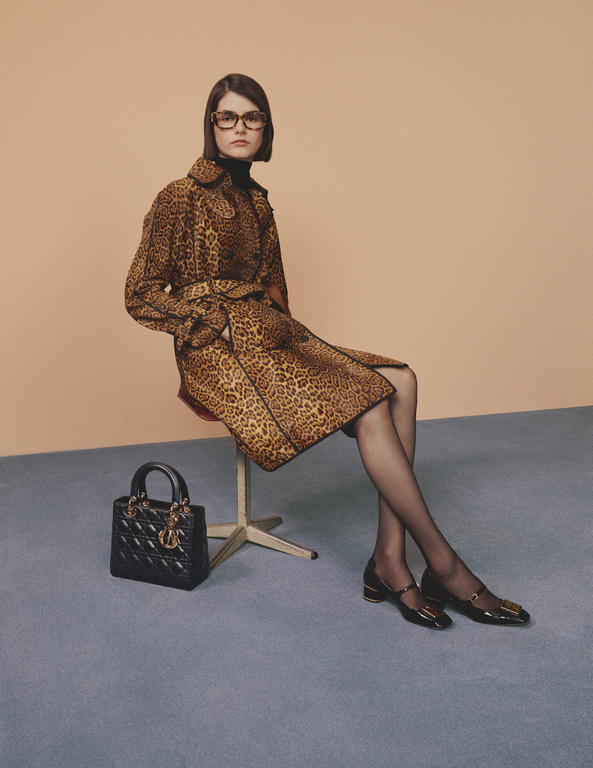 A woman sits in a studio wearing a leopard print coat, black turtleneck, sheer black stockings, and black Mary Jane shoes with a handbag.