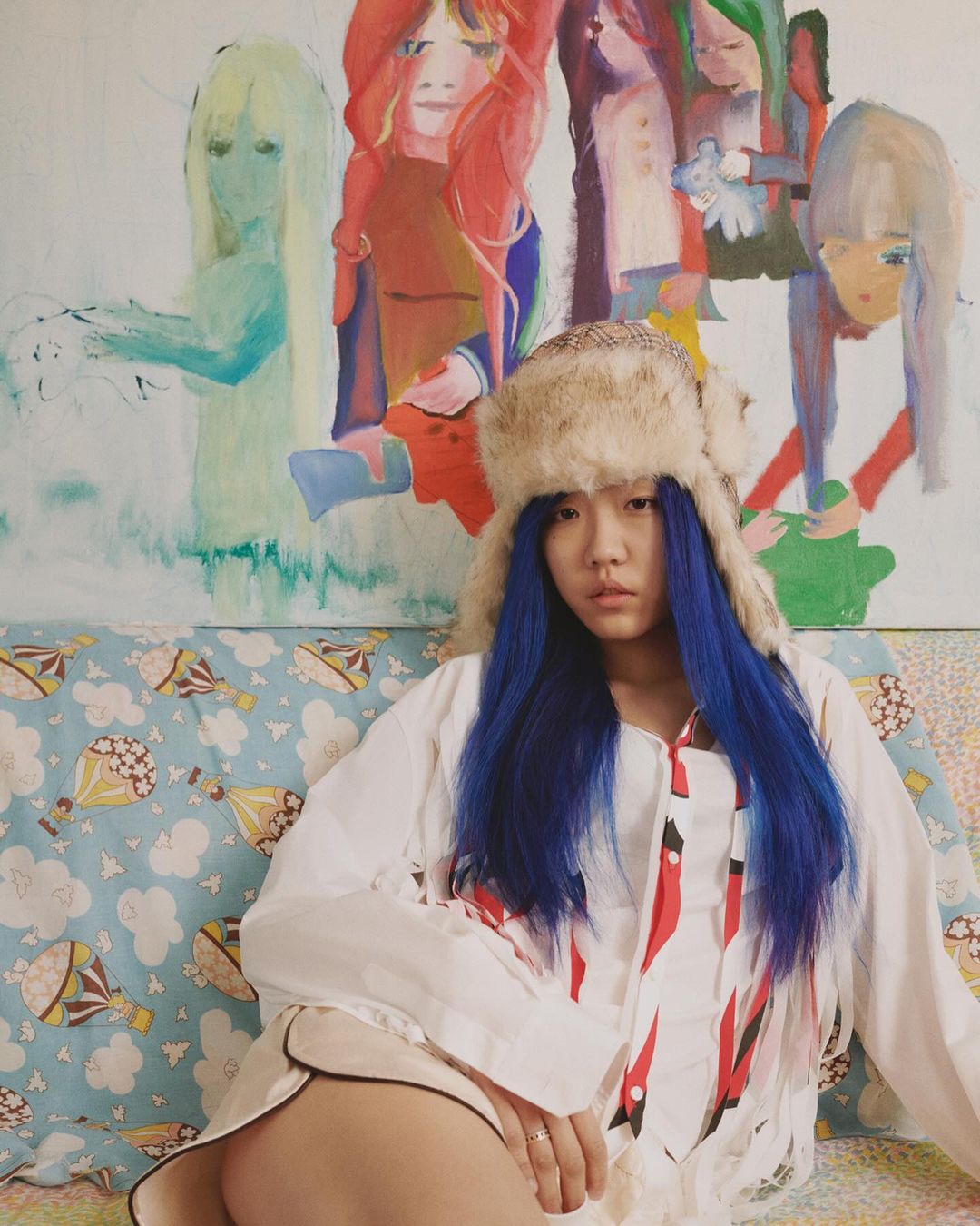 Young woman with blue hair and a fur hat sits on a patterned couch, posing in front of a colorful painting of figures.