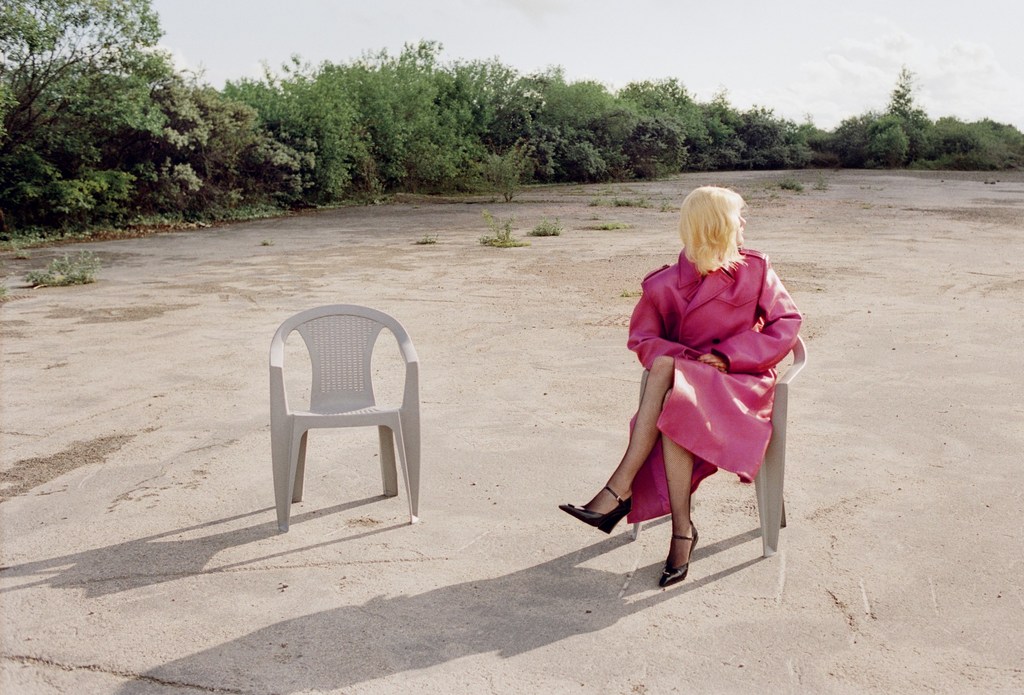 Woman in pink coat sits in chair in outdoor lot, another chair sits nearby, with vegetation and sky in the background.