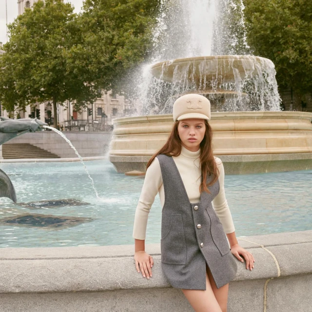 A young woman poses in front of the Trafalgar Square fountain in London, wearing a gray jumper dress and cream-colored turtleneck and hat.