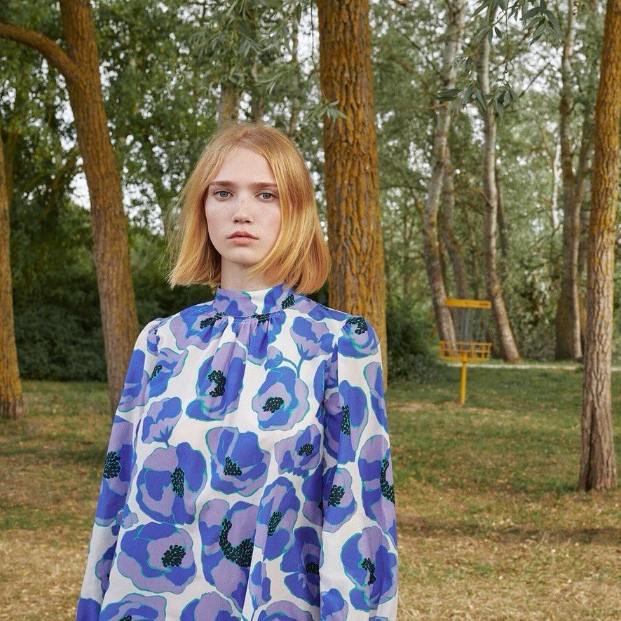 Young woman with short blonde hair wearing a floral patterned blouse poses in a sunny wooded area on a bright day.