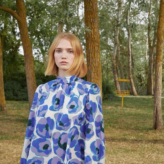 Young woman with short blonde hair wearing a floral patterned blouse poses in a sunny wooded area on a bright day.
