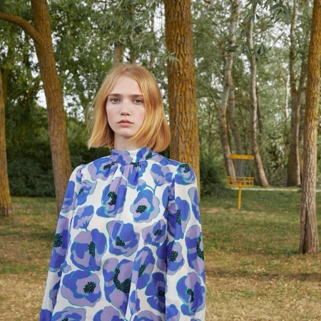 Young woman with short blonde hair wearing a floral patterned blouse poses in a sunny wooded area on a bright day.