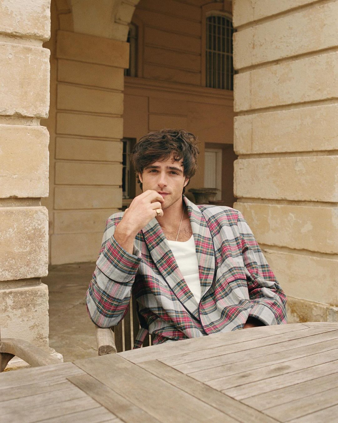 Jacob Elordi sits at a table outdoors, wearing a plaid jacket and white top, in a portrait shot with stone architecture.