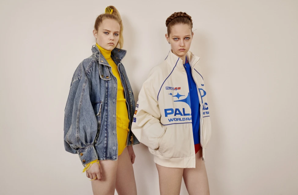 Two young women pose indoors wearing retro-style racing jackets, one denim, one white. Bright yellow tops and bracelets add pops of color.
