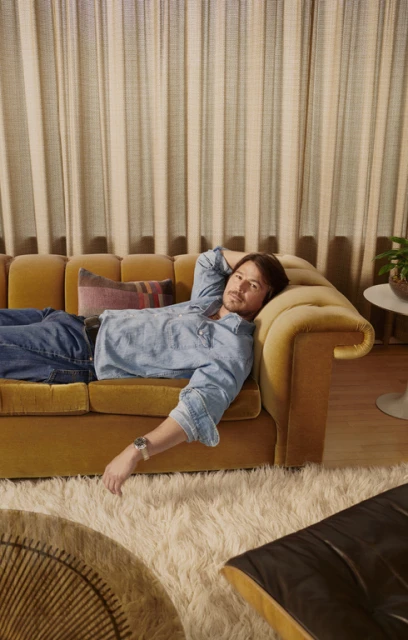 Man lounges on a mustard-yellow velvet sofa with a plaid pillow in a living room with shag carpet and cream-colored curtains.