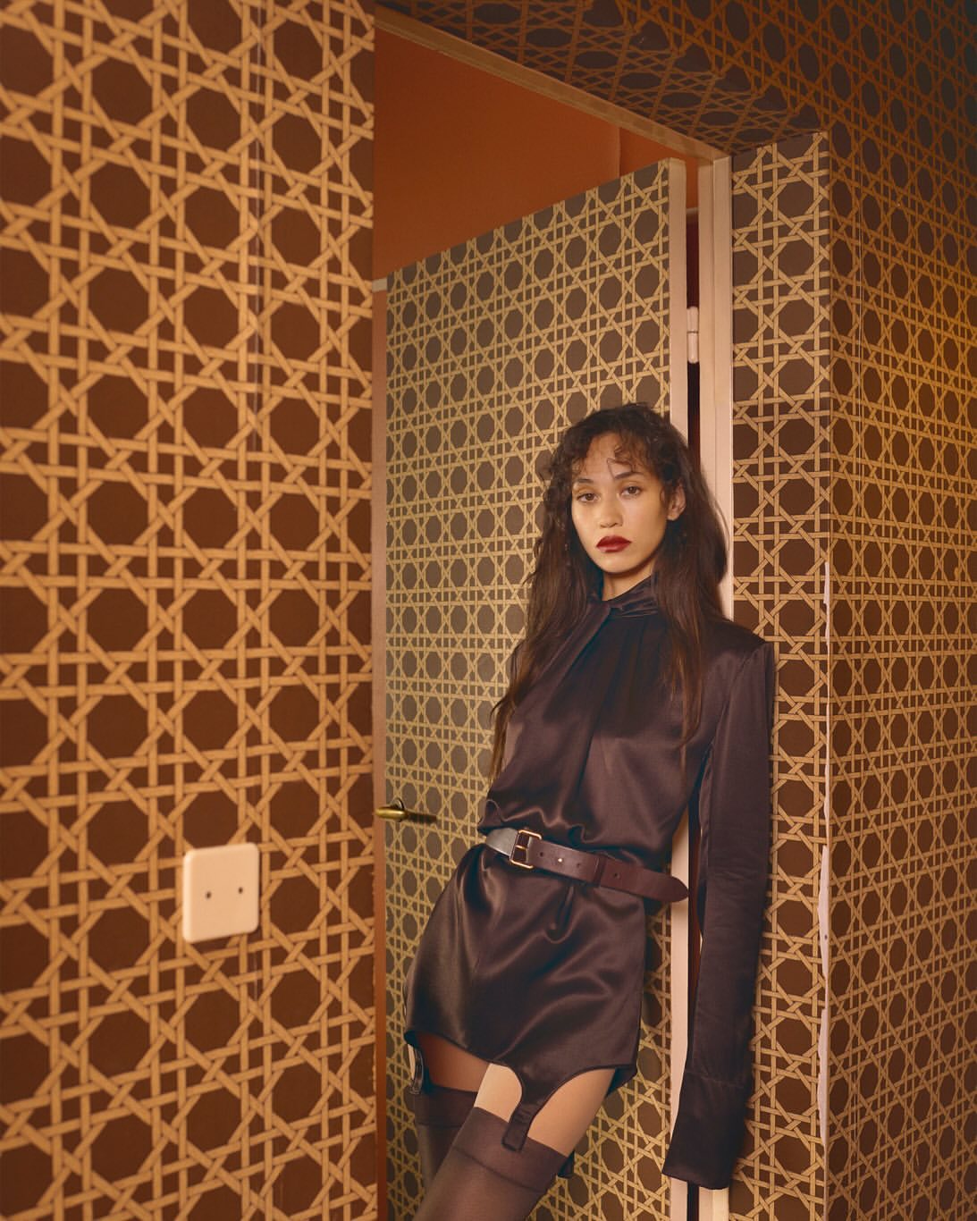 A woman in a dark brown outfit poses in front of a retro patterned wall with a doorway. The image evokes a vintage editorial feel.