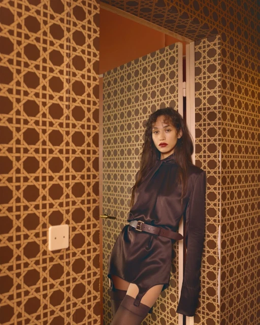 A woman in a dark brown outfit poses in front of a retro patterned wall with a doorway. The image evokes a vintage editorial feel.