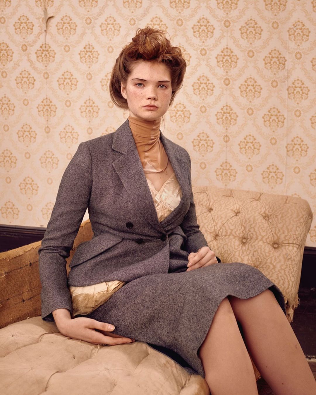 Indoor portrait of a woman with auburn hair wearing a grey business suit and tan turtleneck sweater, posing in front of patterned wallpaper.
